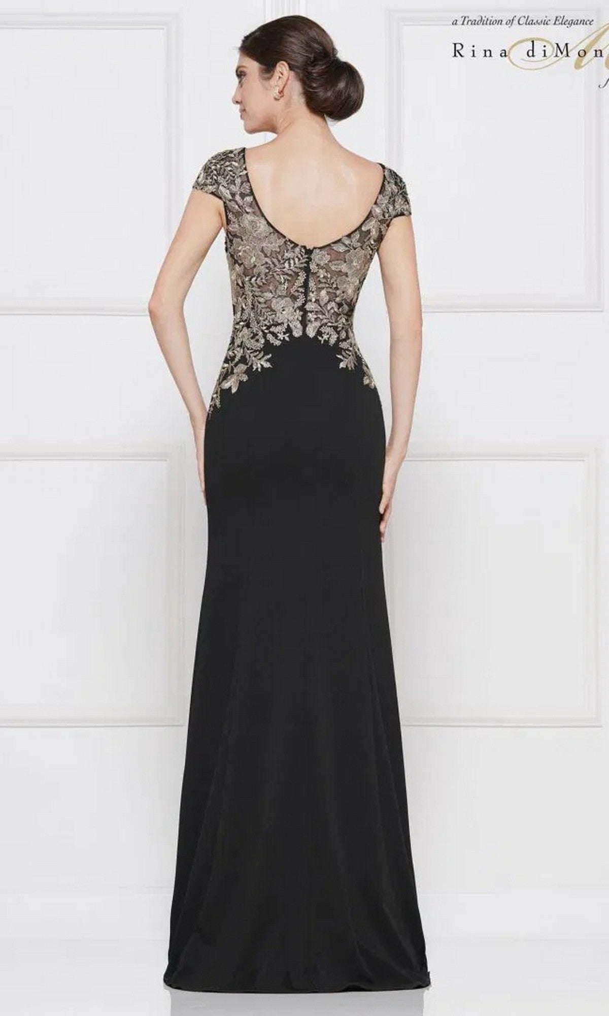 A woman stands before a white wall in the Rina Di Montella RD2652, a floor-length black formal dress with a sheath skirt and metallic gold-embroidered floral detail on the upper back.